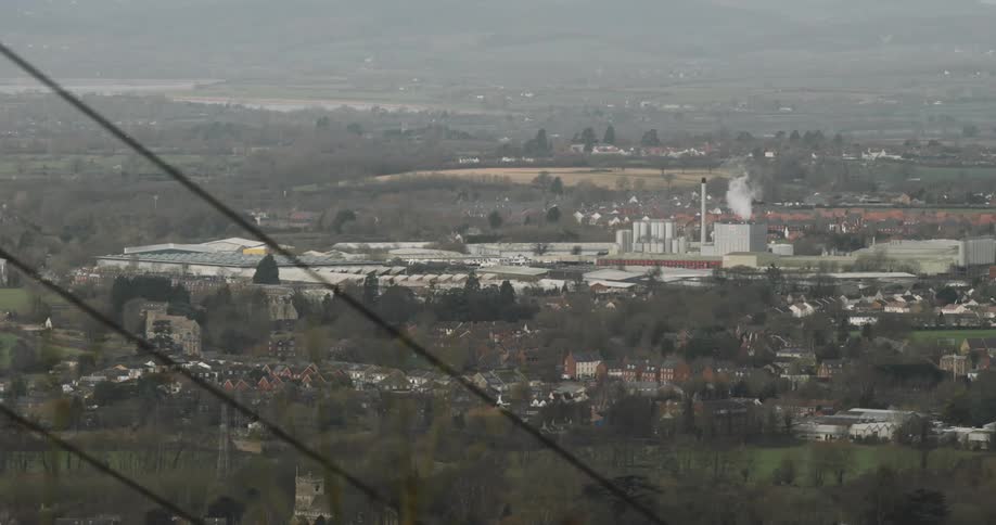 Smoke rises from a factory situated in a rural town located between waterways on a foggy day