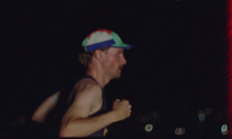 A man in a cap and black tank top is running quickly in a place at night