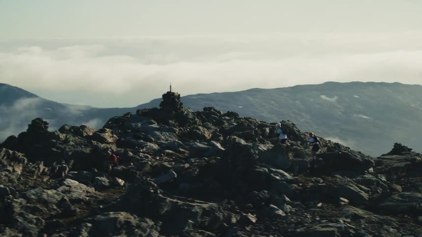 Two backpackers race up a rugged rocky slope to stand at a peak overlooking a valley on a foggy day