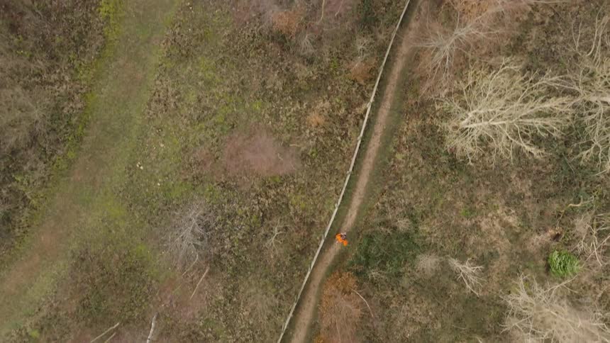 A person in orange clothes is running on a dirt path in an area with grass and dry trees seen from above