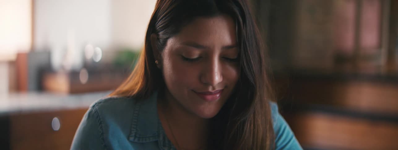 A teenage girl sits inside a house and stares down while smiling