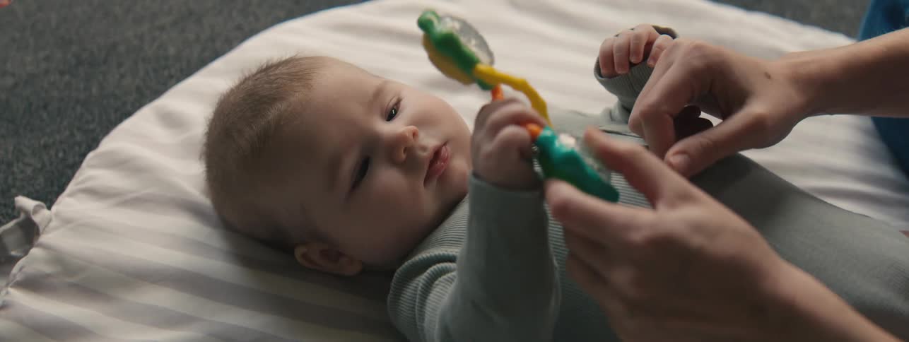 An adult hand shakes a brightly colored rattle as a young baby holds it