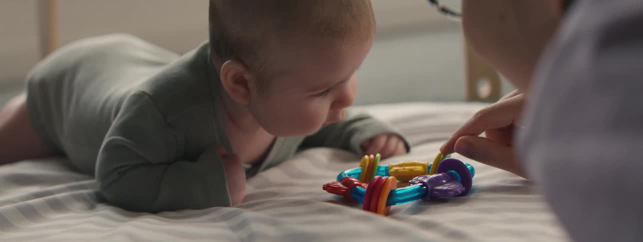 Father shakes toy in front of baby lying on a cushion at home