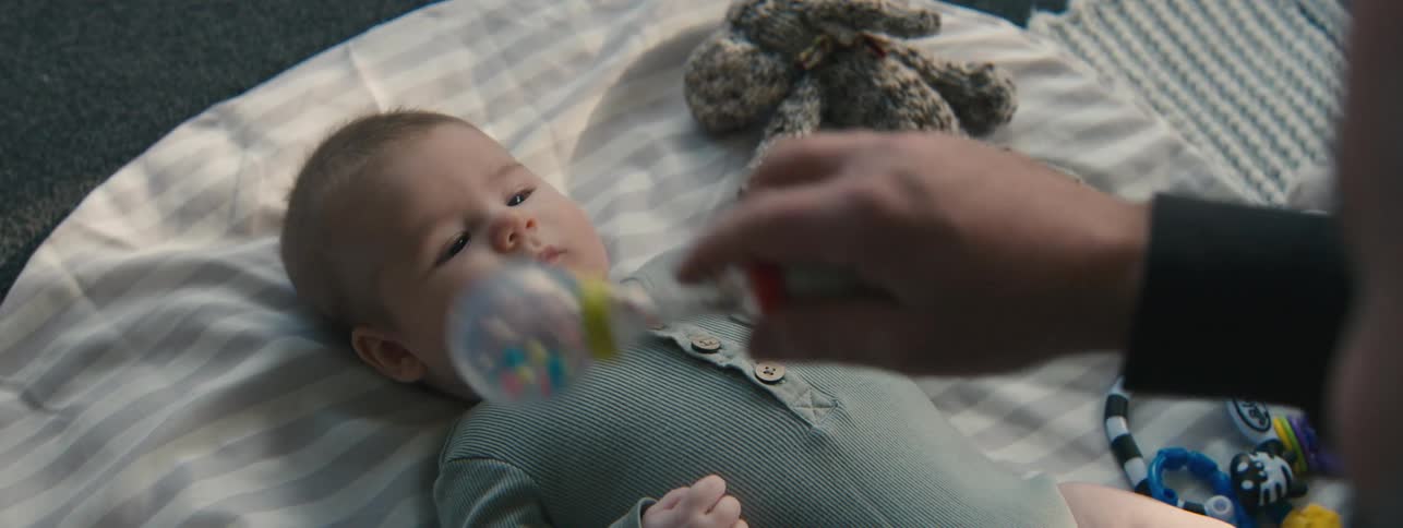 A father shakes a rattle above his baby son who is lying on a blanket