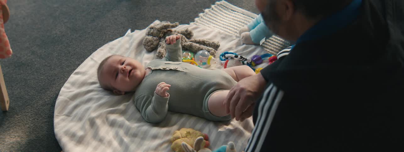 An older man rubs a babys knees while the baby lies on a blanket on the floor