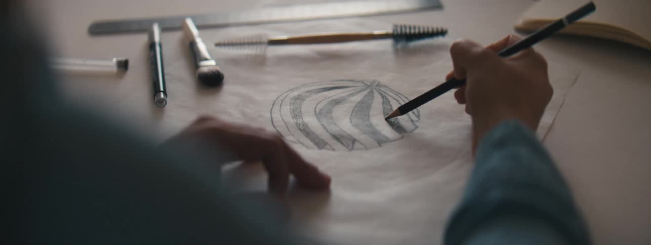 A human hand using a black pencil to complete a drawing of a ball made on paper