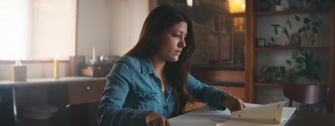 A closeup of a woman in a denim shirt working on a drawing at a desk in a warmly lit room