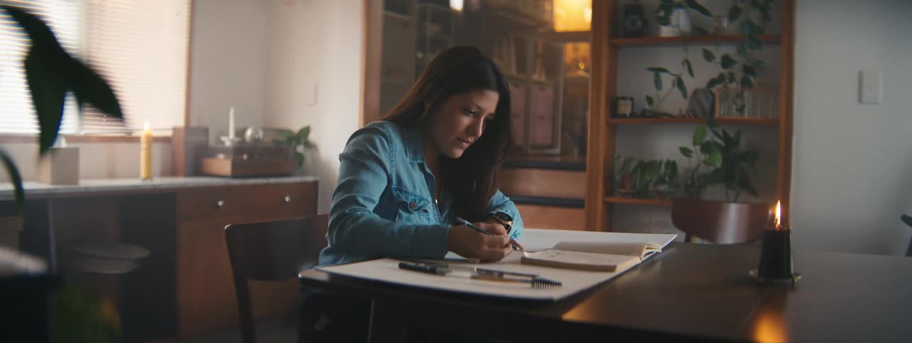 A closeup of a woman in a denim shirt working on a drawing at a desk in a warmly lit room