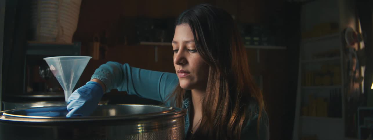 A teenage girl stirs a substance in a large drum out in a garage