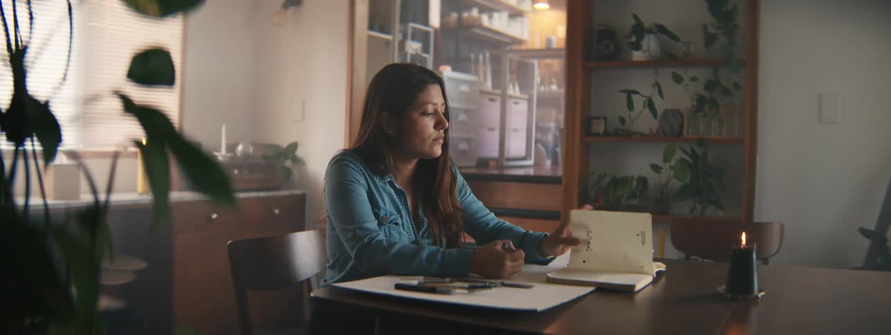 Woman sitting at a table in a room drawing and turning the pages of a book in front of her