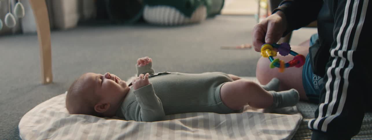 An adults hand holds a colorful baby rattle above a baby laying on a gray blanket