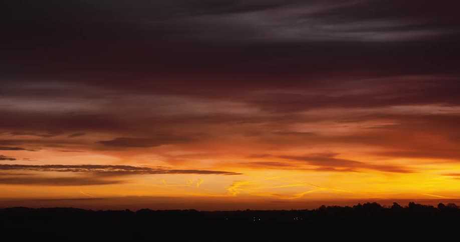 A fiery sunset casts warm hues across the sky with a dark silhouette of trees on the distant horizon