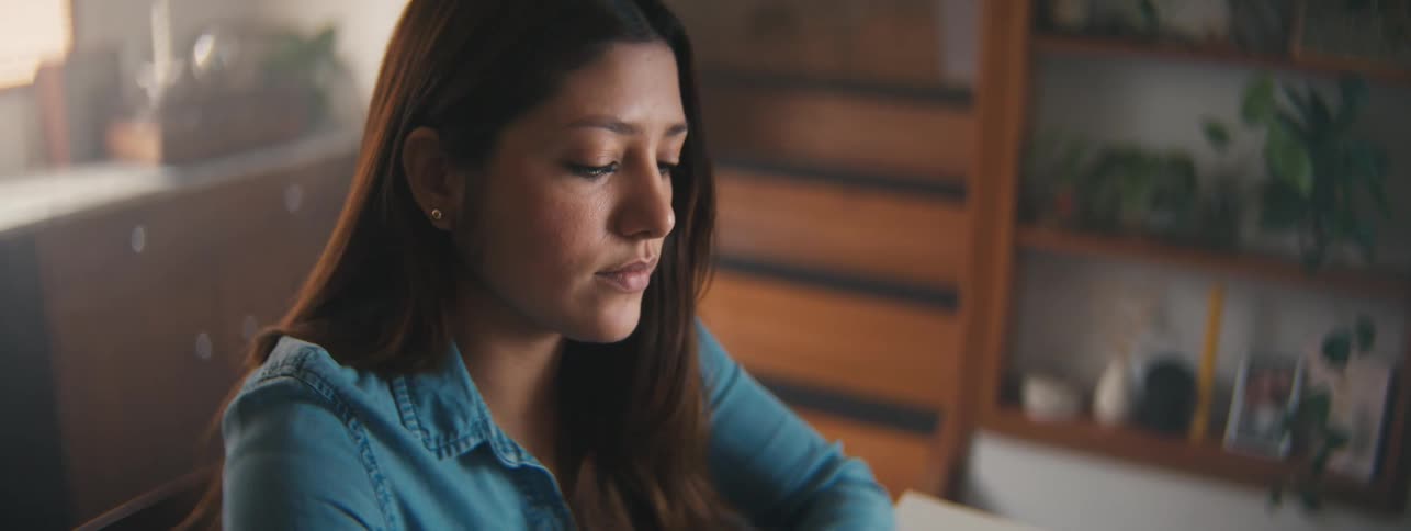 A teenage girl wearing a denim shirt sits inside a house while thinking and looking downward