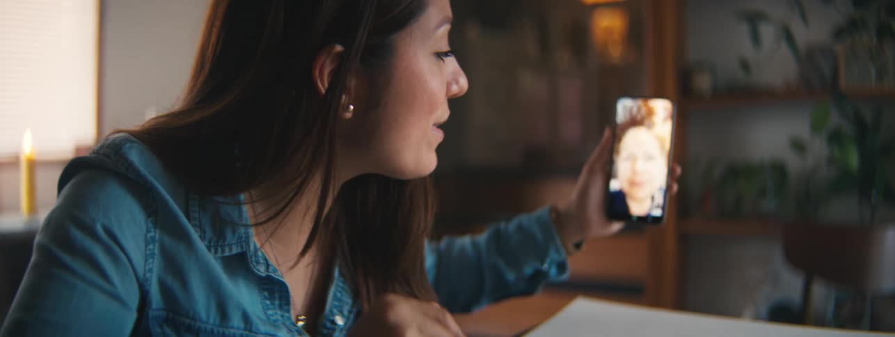 A teenage girl sits at a table talking and waving to an older woman on a smartphone