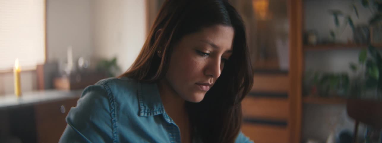 A closeup of a woman in a denim shirt working at a desk in a warmly lit room