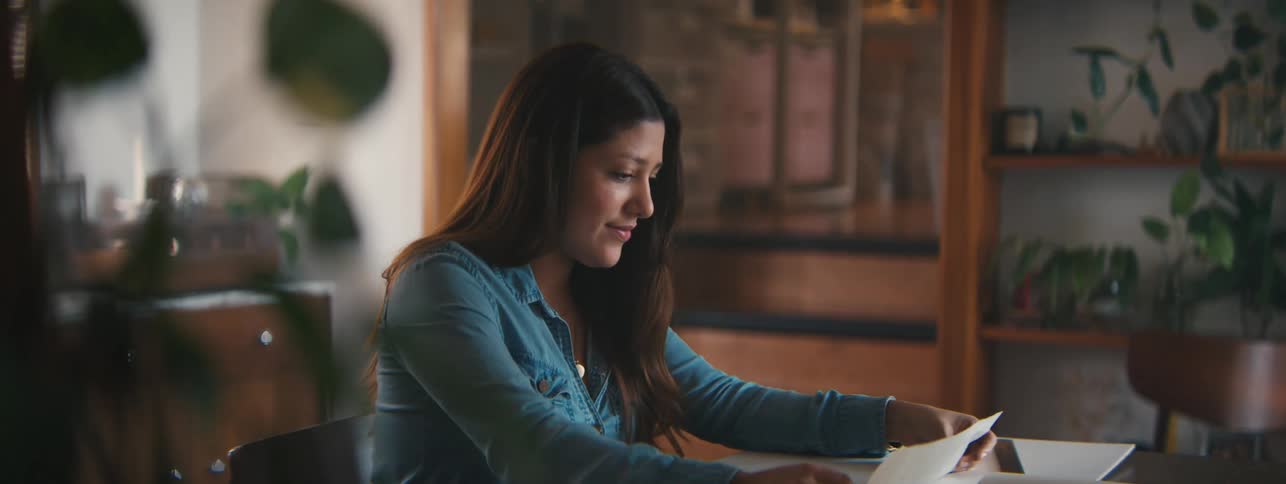 A closeup of a woman in a denim shirt working on a drawing at a desk in a warmly lit room
