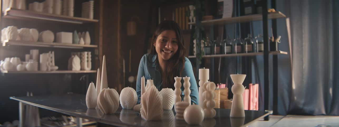 Female candle maker smiles while sitting at a table with candles in her workshop