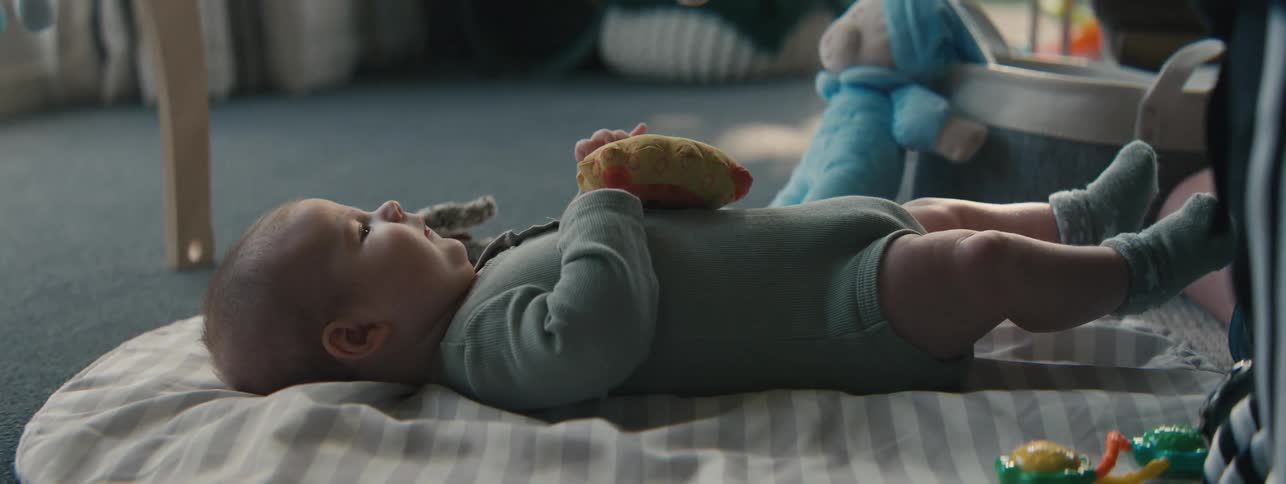 A baby lies on a play mat with toys around them watching a hand waving a stuffed animal