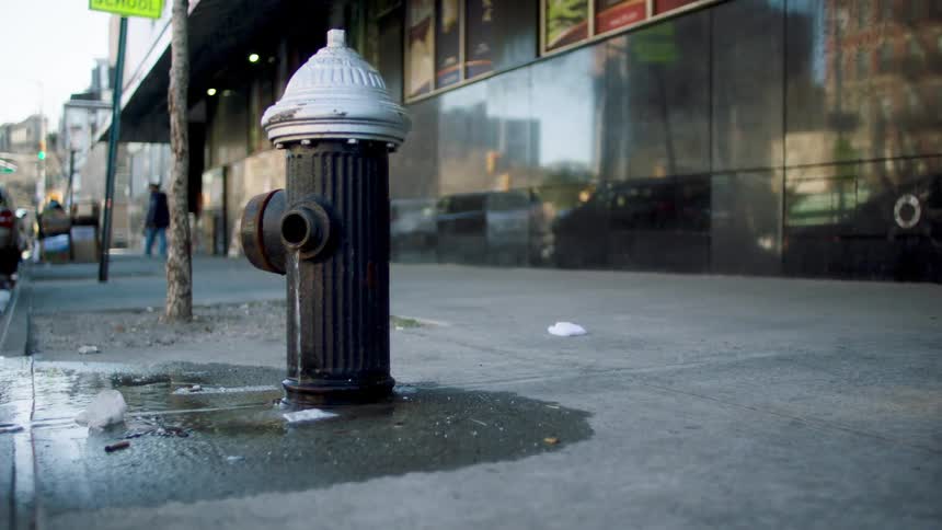A city street with a fire hydrant on the sidewalk water trickling down and pedestrians in the background