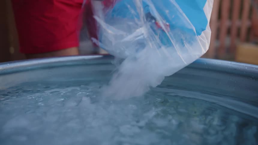 A man in a backyard preparing an ice bath by pouring ice from a plastic bag into a metallic basin with water