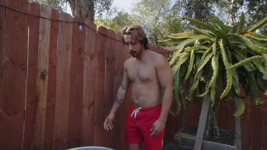 A shirtless muscular young man prepares to take an outdoors bath on a metal tub in the backyard