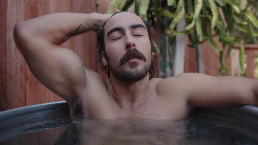 A sad looking man with a sleeve tattoo bathes in a tub in a backyard