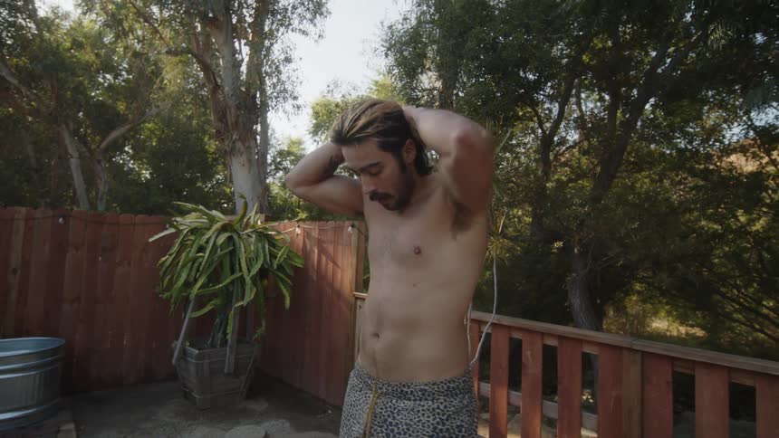 Shirtless man in a backyard surrounded by trees stretching his body