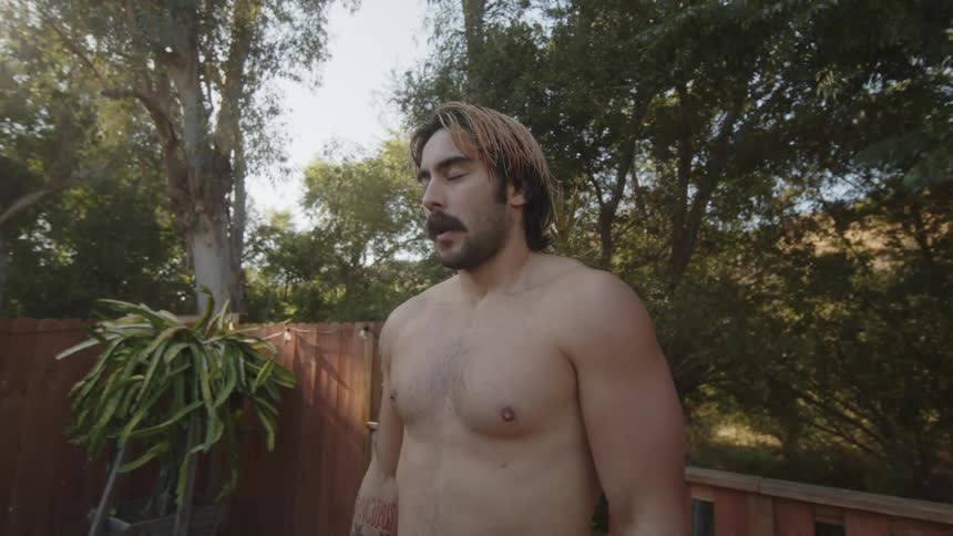 Bearded man with no shirt in backyard surrounded by trees taking deep breaths