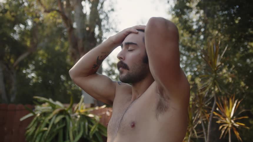 Shirtless man tossing his hair back while standing in a treelined environment