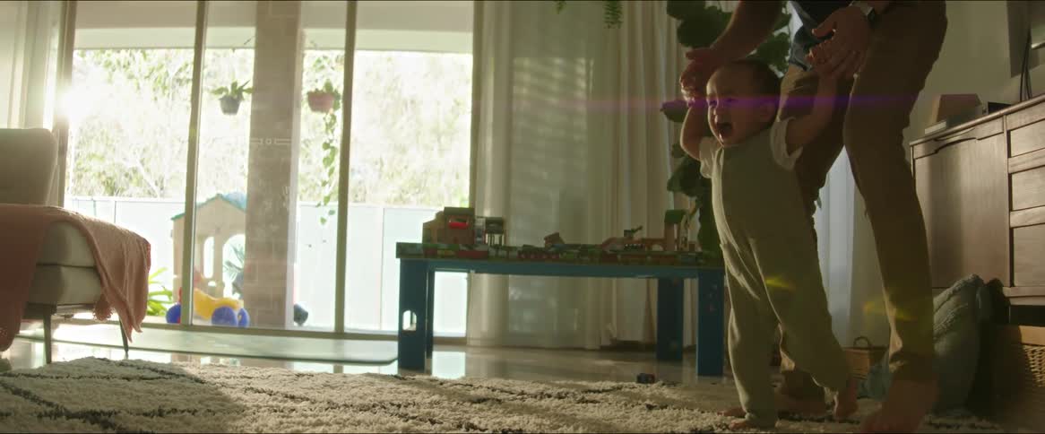 A father helps his screaming and crying son walk across a rug in a living room