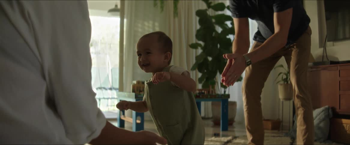 Parents clap and encourage their toddler son to walk across a living room floor