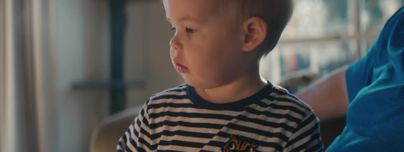 A toddler bounces next to an adult on a couch while he holds a portable speaker