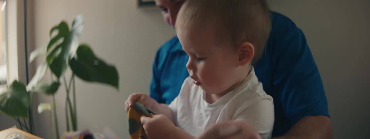 A little blond boy in a white shirt who is on the lap of an adult who is fiddling with a small sock that is in his hands