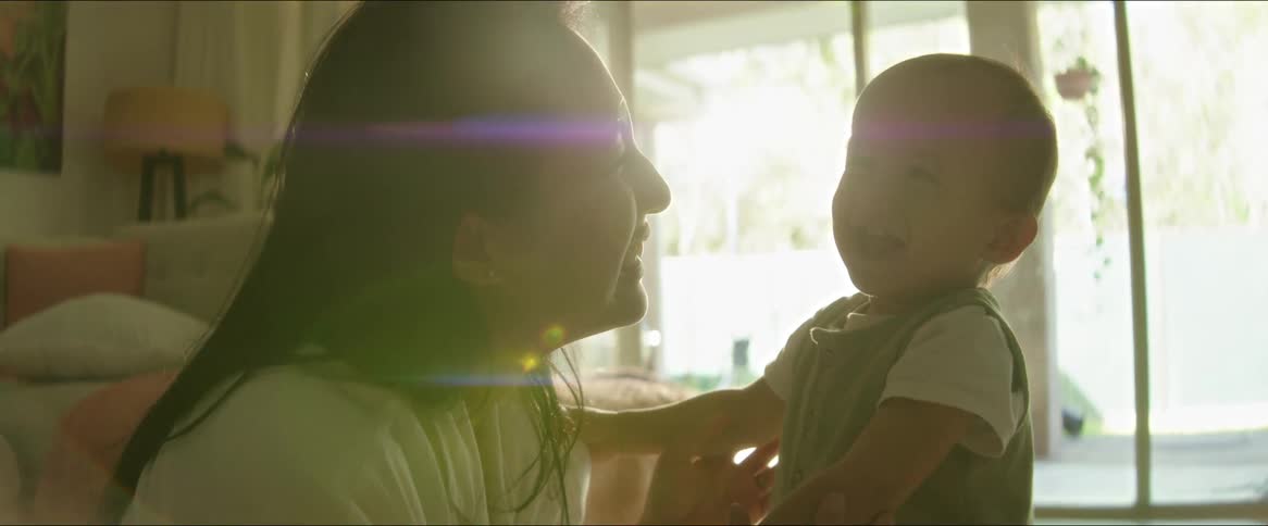 A mother and her toddler son laugh and smile as the sun shines into the living room