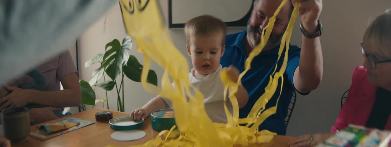 A family sitting around a table watches a toddler grab yellow strips from a minion toy