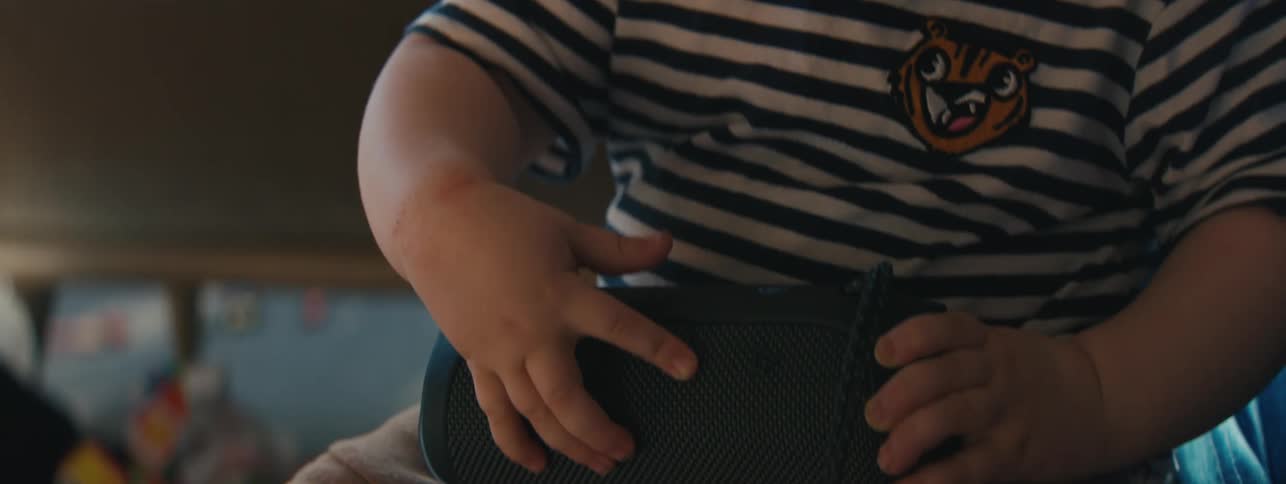 A toddler sits next to an adult on a couch while he fiddles with a portable speaker