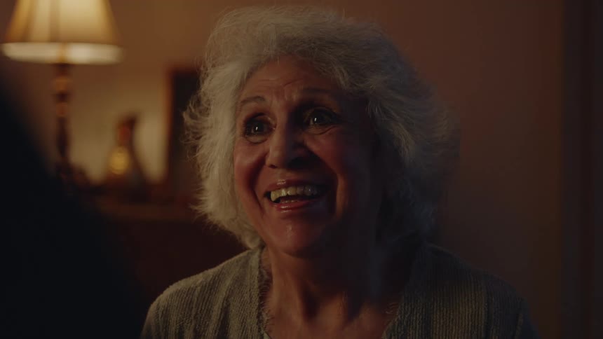 An elderly woman in a living room stands across from someone else and smiles