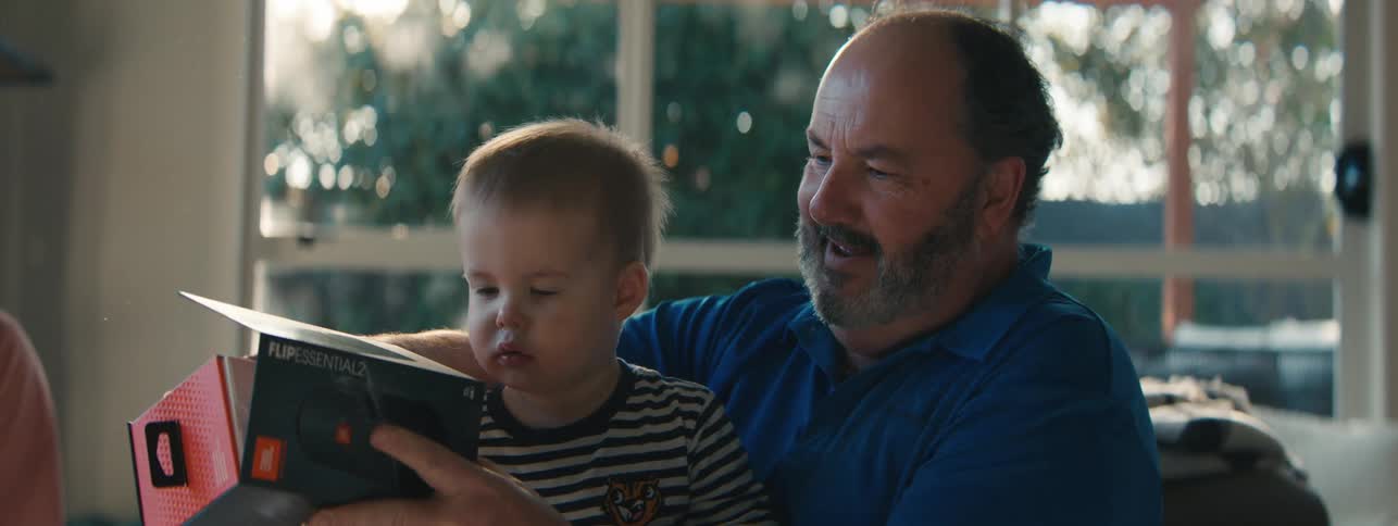 Grandfather sitting in the living room holds his grandson as they examine the contents of a box