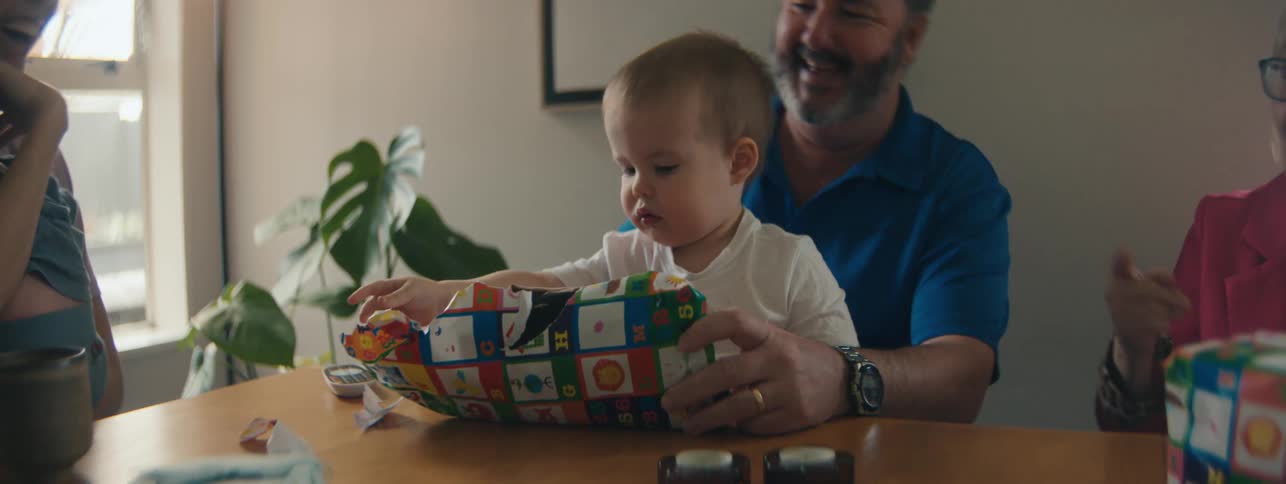 Small child on grandfathers lap opening a present and with a family sitting at the table around
