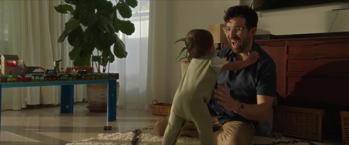 A mother and father laughing seeing their toddler walk across a rug to his father who picks him up and tosses him into the air