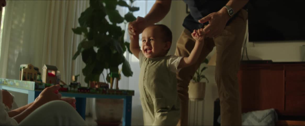 A little boy is walking on a carpet with his arms held by an adult person towards a woman who is in front of him clapping and smiling and then she hugs him
