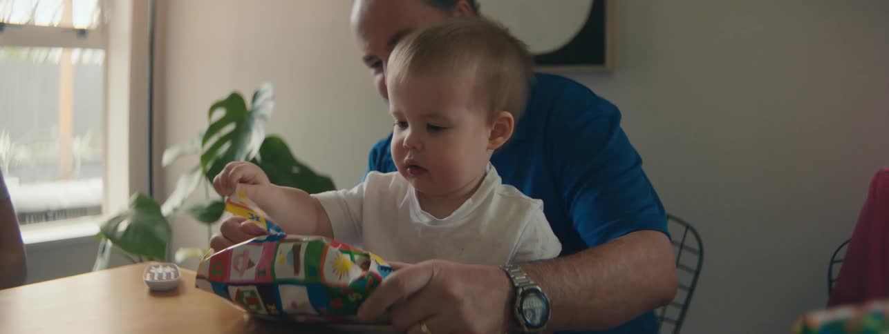 Small child sitting on fathers lap opening a gift at table with family around