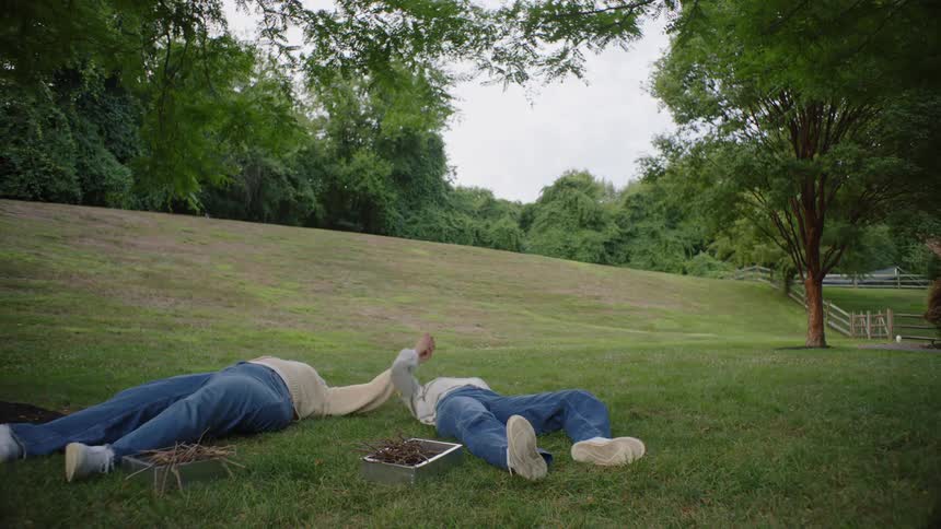 Two people lying in a field near a fenced pasture hold hands