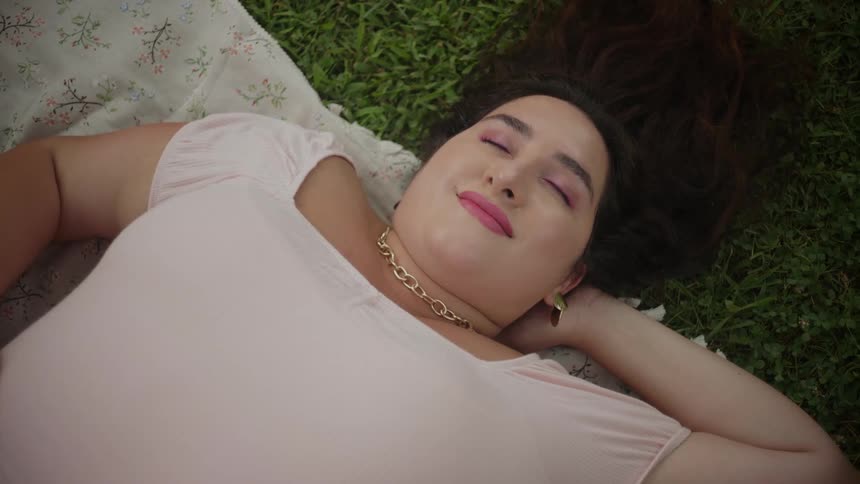 A woman lies on a blanket on grass with her eyes closed and a smile on her face