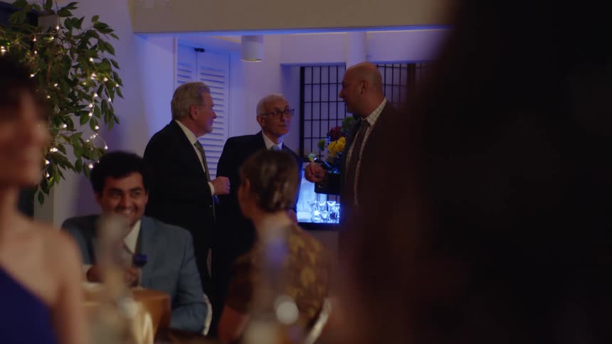 Older men stand and talk while other people sit at dinner tables during a party