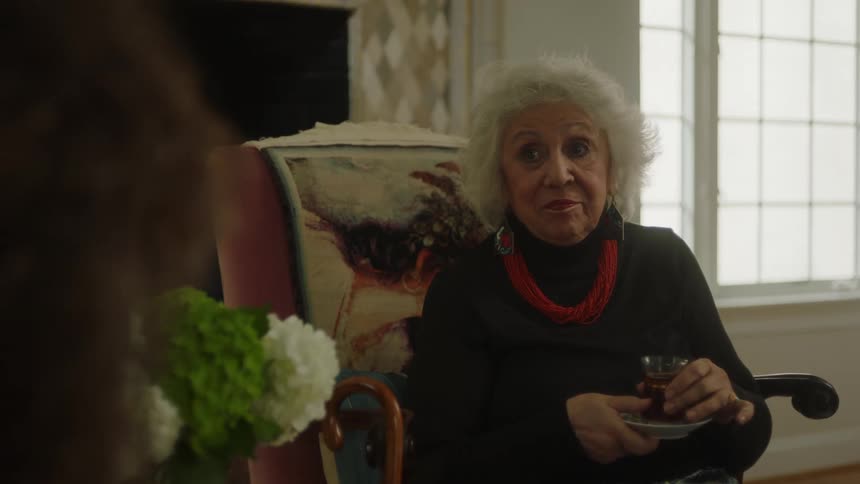An older woman sitting in a chair and holding a drink looks at another woman and nods her head