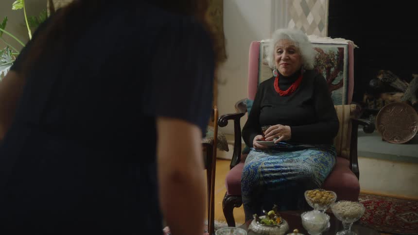 A woman walks into a room and sits down as one woman looks on with a smile as another woman looks angry