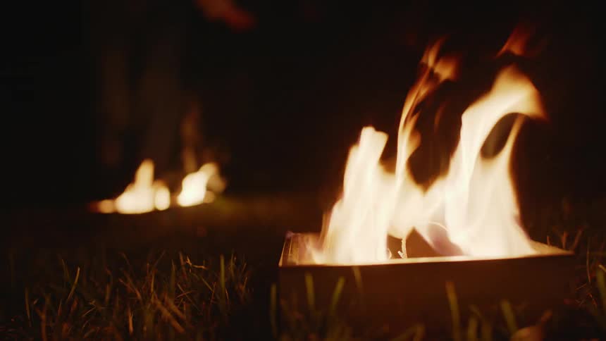 Person wearing jeans jumps over fires burning in boxes on the ground at night