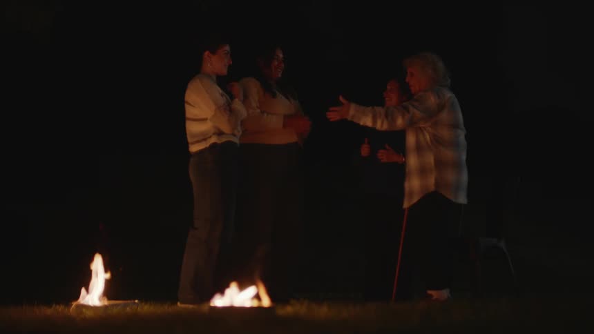 Group of four women of different ages in front of bonfires with one of them jumping on top while in a natural setting at night