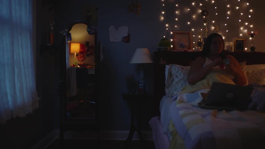 A woman sits in bed with her laptop while eating a bag of chips